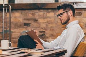 notando il suo idee. lato Visualizza di bello giovane uomo nel bicchieri scrittura nel il suo Nota tampone mentre seduta a il suo Lavorando posto foto