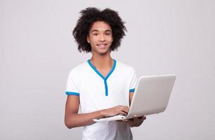 adolescente con il computer portatile. allegro africano adolescente Lavorando su il computer portatile e sorridente a telecamera mentre in piedi isolato su grigio sfondo foto