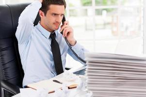 occupato ufficio vita. frustrato giovane uomo nel vestito formale parlando su il telefono e Tenere mano nel capelli mentre seduta a il suo Lavorando posto foto