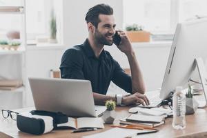 bene attività commerciale parlare. bello giovane uomo parlando su il mobile Telefono mentre seduta a il suo Lavorando posto nel creativo ufficio foto