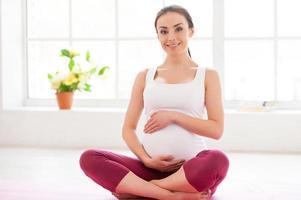 conservazione calma per sua nascituro bambino. bellissimo incinta donna seduta nel loto posizione su esercizio stuoia e sorridente foto