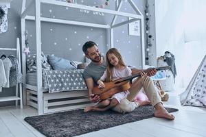 lui sempre crede nel suo. giovane padre insegnamento il suo poco figlia per giocare chitarra e sorridente mentre seduta su il pavimento nel Camera da letto foto