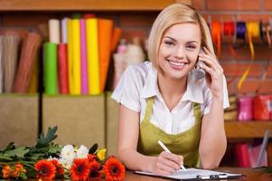 bellissimo fioraio a opera. bellissimo giovane femmina fioraio scrittura qualcosa a Nota tampone e assunzione su il Telefono mentre in piedi a sua Lavorando posto foto