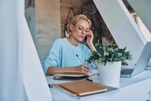 bellissimo giovane donna scrittura qualcosa nel Nota tampone e parlando su mobile Telefono mentre Lavorando nel ufficio foto