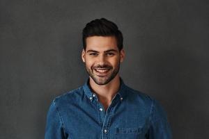 contento uomo. bello giovane uomo nel denim camicia guardare a telecamera e sorridente mentre in piedi contro grigio sfondo foto