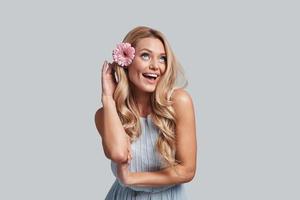 fresco e carino. attraente giovane donna con un' fiore nel capelli conservazione braccia attraversato e sorridente mentre in piedi contro grigio sfondo foto