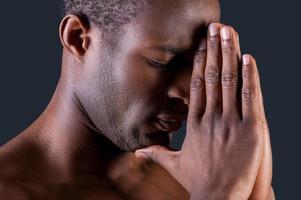 preghiere uomo. lato Visualizza di giovane africano uomo preghiere mentre Tenere mani stretto vicino e contro grigio sfondo foto