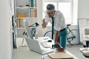 così occupato. bene guardare giovane uomo potabile caffè e sorridente mentre in piedi nel il ufficio foto