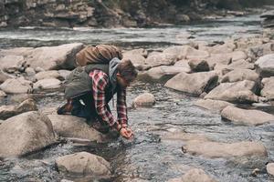 bisogno rinfresco. bello giovane moderno uomo potabile acqua a partire dal il fiume mentre escursioni a piedi nel montagne foto