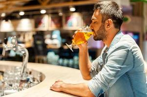 godendo fresco birra. lato Visualizza di giovane uomo potabile birra e conservazione occhi chiuso mentre seduta a il bar contatore foto