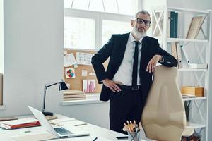 fiducioso maturo uomo nel pieno completo da uomo guardare a telecamera mentre Lavorando nel moderno ufficio foto