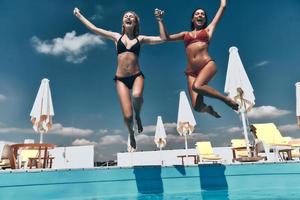 vero la gioia. pieno lunghezza di Due attraente giovane donne nel bikini Tenere mani e mentre salto nel il piscina insieme foto