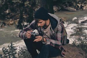 sempre trasporto telecamera. giovane moderno uomo con zaino Tenere un' foto telecamera mentre seduta nel il boschi con fiume nel sfondo
