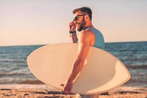 onda amante. bello giovane uomo Tenere skimboard e regolazione occhiali mentre a piedi lungo il spiaggia foto