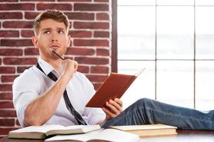 perso nel pensieri. riflessivo giovane uomo nel camicia e cravatta scrittura qualcosa nel Nota tampone e guardare lontano mentre seduta a il suo Lavorando posto foto