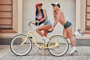 ragazze avendo divertimento. lato Visualizza di contento giovane donna equitazione su bicicletta mentre sua femmina amico pattinando dietro a sua foto