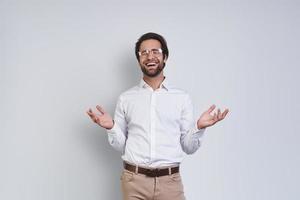 bello giovane sorridente uomo nel bianca camicia Esprimere a gesti mentre in piedi contro grigio sfondo foto