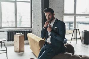 pensiero di... bello giovane uomo nel pieno completo da uomo regolazione il suo camicia mentre pendente su il divano foto