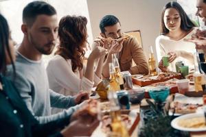 così gustoso gruppo di giovane persone nel casuale indossare mangiare Pizza mentre avendo un' cena festa in casa foto