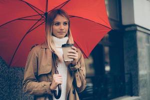 riscaldamento su con caldo caffè. attraente giovane donna trasporto ombrello e caffè tazza mentre in piedi su il strada foto