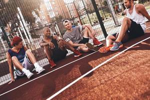 migliore gli amici. gruppo di giovane uomini nel gli sport capi di abbigliamento sorridente mentre seduta su il pallacanestro campo all'aperto foto