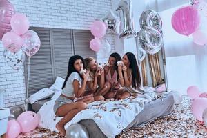 così gusto quattro bellissimo giovane donne nel pigiama mangiare torta mentre avendo un' sonno festa nel il Camera da letto con palloncini tutti al di sopra di il posto foto