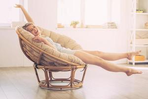 godendo spensierato tempo a casa. allegro giovane donna conservazione occhi chiuso e allungamento su mani mentre seduta nel grande confortevole sedia a casa foto