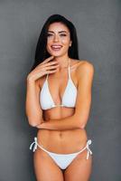 fresco e Bellissima. attraente giovane sorridente donna nel bianca bikini in posa contro grigio sfondo foto