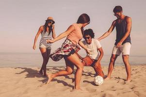 avendo divertimento con gli amici. gruppo di allegro giovane persone giocando con calcio palla su il spiaggia con mare nel il sfondo foto