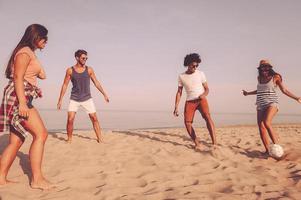 godendo tempo con gli amici. gruppo di allegro giovane persone giocando con calcio palla su il spiaggia con mare nel il sfondo foto