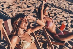 vero la gioia. superiore Visualizza di attraente giovane donne sorridente e prendere il sole mentre riposo nel all'aperto sedie su il spiaggia foto