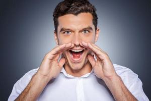 annunciando grande notizia. contento giovane uomo nel bianca camicia urlando mentre guardare a telecamera e in piedi contro grigio sfondo foto