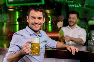 uomo nel birra pub. bello giovane uomo Tenere un' birra boccale e sorridente mentre barista scrosciante birra su il sfondo foto
