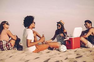 spiaggia volta. allegro giovane persone la spesa simpatico tempo insieme mentre seduta su il spiaggia e potabile birra foto