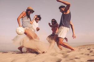 avendo grande divertimento su spiaggia. gruppo di allegro giovane persone giocando con calcio palla su il spiaggia foto
