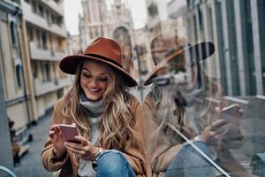 soggiorno collegato. attraente giovane donna nel cappello e cappotto utilizzando sua inteligente Telefono mentre la spesa spensierato tempo nel il città foto