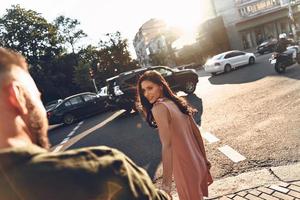 bellissimo giovane donna guardare a sua fidanzato e Tenere il suo mano mentre a piedi insieme attraverso il città strada foto