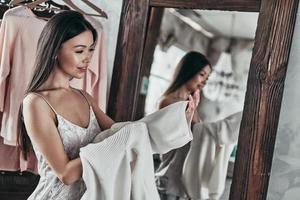 Perfetto camicetta. attraente giovane donna assunzione su il camicetta e sorridente mentre in piedi nel davanti di il specchio a casa foto