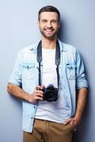 fiducioso fotografo. bello giovane uomo Tenere digitale telecamera e sorridente mentre in piedi contro grigio sfondo foto