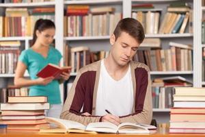 preparazione per il esami. riflessivo giovane uomo seduta a il biblioteca scrivania e scrittura qualcosa nel il suo Nota tampone mentre giovane donna lettura un' libro su il sfondo foto