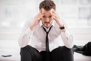 sensazione stanco e depresso. frustrato giovane uomo nel camicia e cravatta Tenere testa nel mani mentre seduta su il letto foto