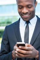 digitando attività commerciale Messaggio. allegro giovane africano uomo nel vestito formale Tenere mobile Telefono e sorridente mentre in piedi all'aperto foto