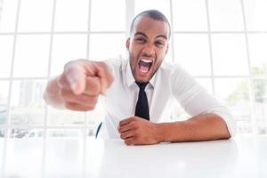 furioso capo. furioso giovane africano uomo nel camicia e cravatta puntamento voi e urlando mentre seduta a il suo Lavorando posto foto