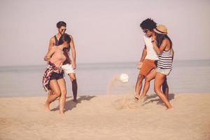 la spesa tempo con migliore gli amici. gruppo di allegro giovane persone giocando con calcio palla su il spiaggia con mare nel il sfondo foto