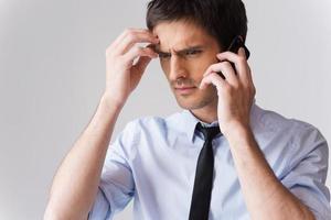 veramente cattivo notizia. frustrato giovane uomo nel camicia e cravatta parlando su il mobile Telefono e toccante testa con mano mentre in piedi contro grigio sfondo foto