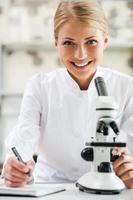 giovane e fiducioso scienziato. sorridente giovane femmina scienziato guardare a telecamera e scrittura nel Nota tampone mentre seduta a sua Lavorando posto foto