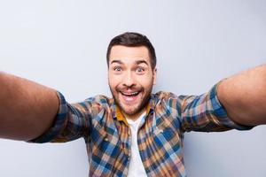 io amore autoscatto bello giovane uomo nel camicia Tenere telecamera e fabbricazione autoscatto e sorridente mentre in piedi contro grigio sfondo foto