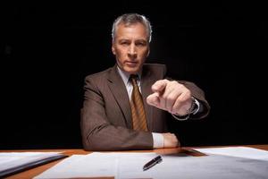 io scegliere voi grave anziano uomo nel vestito formale seduta a il suo Lavorando posto e puntamento voi mentre isolato su nero sfondo foto