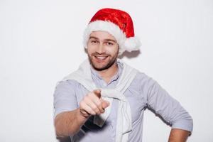 allegro Natale per voi bello giovane uomo nel Santa cappello puntamento voi e sorridente mentre in piedi contro bianca sfondo foto