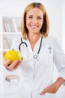 mangiare salutare allegro femmina medico nel bianca uniforme allungamento su mano con verde Mela e sorridente foto